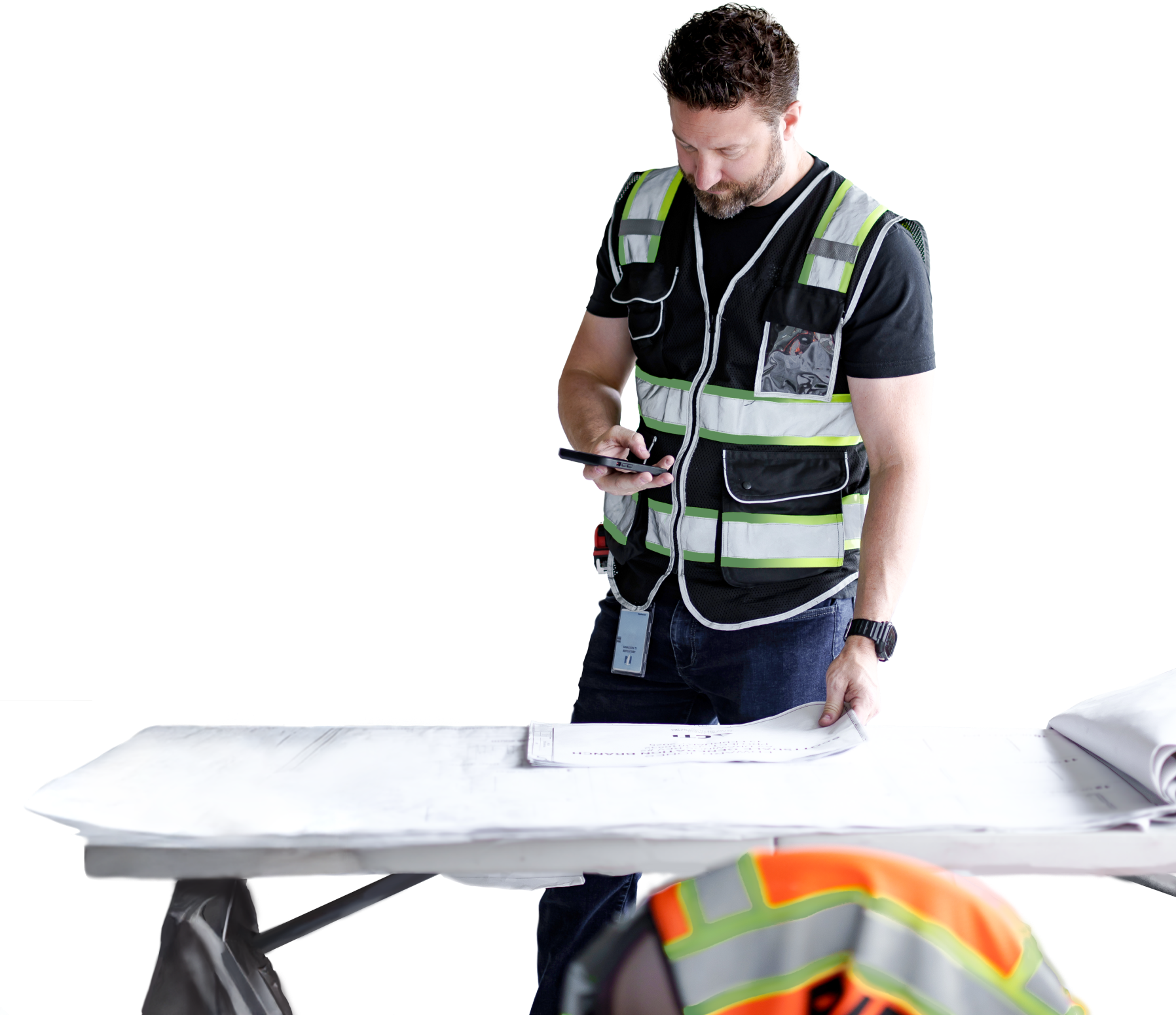 A construction worker in a safety vest looks at a smartphone while standing at a table with blueprints on a commercial construction Arizona site; another worker in a helmet is partially visible in the foreground.