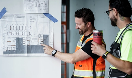 Two men in safety vests discuss a floor plan taped to a wall on a commercial construction job in Arizona; one points at the plan while the other holds a drink cup.