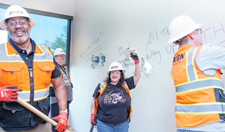 Four construction workers wearing safety gear remove drywall and write on a wall inside a building. One, smiling, holds a piece of torn drywall—showcasing teamwork typical of commercial construction jobs in Arizona.