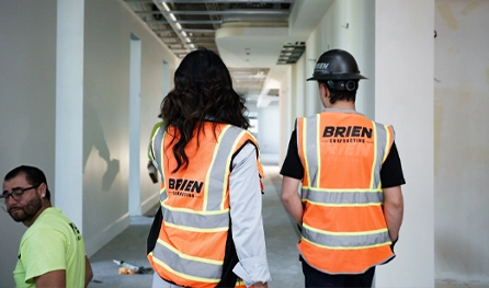 Two people in orange safety vests and hard hats walk down a hallway under construction, showcasing the teamwork involved in commercial construction jobs Arizona, while another person in a yellow shirt sits on the floor.