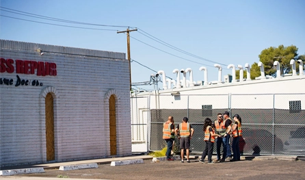 A group of people wearing orange safety vests stand near a chain-link fence outside a building on a sunny day, representing commercial construction jobs Arizona offers.