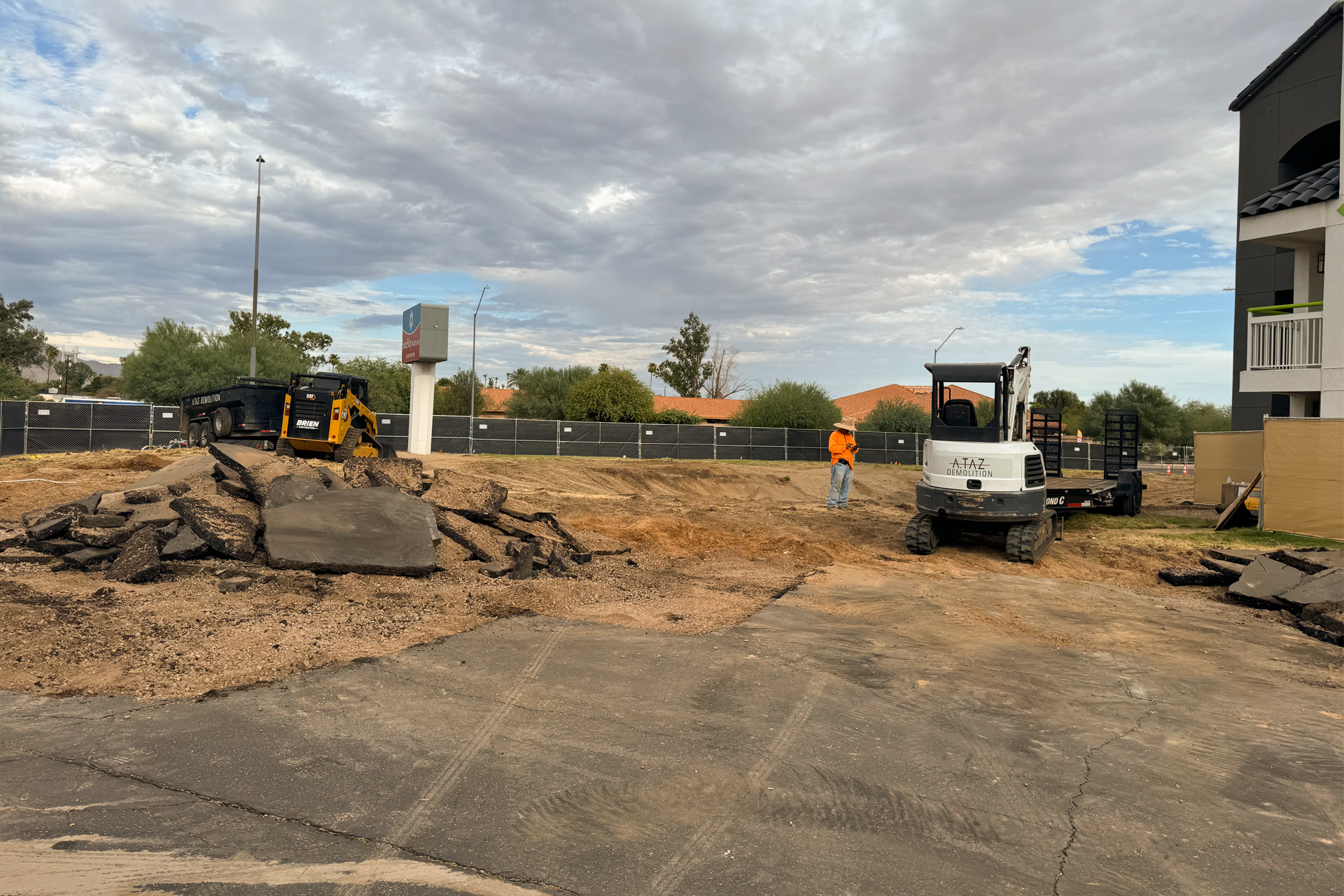 Commercial construction Arizona project in progress: heavy machinery breaks and removes asphalt while a worker in an orange shirt stands near a white excavator.